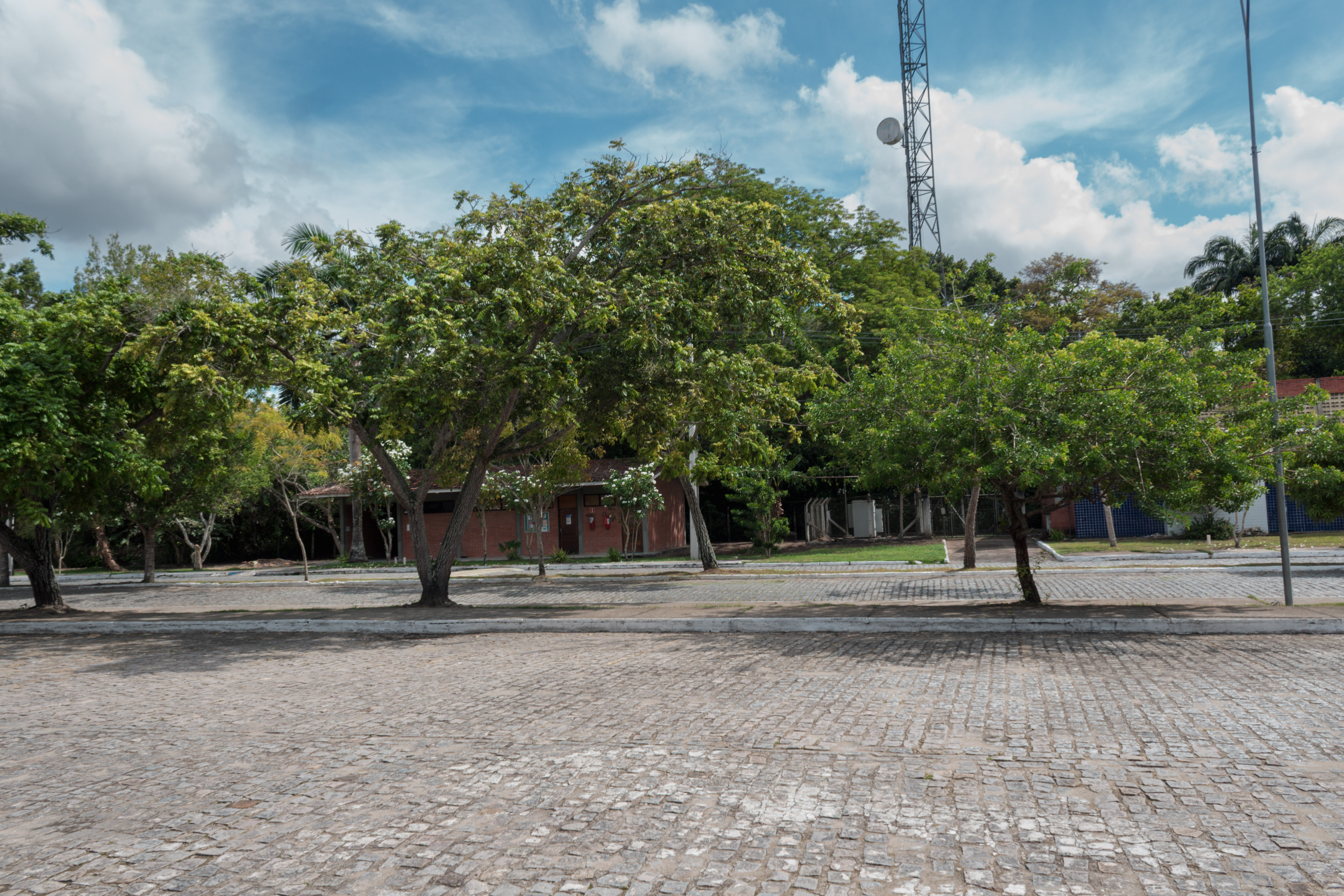 Praça de pedras sombreada por árvores com uma torre de telecomunicações ao fundo